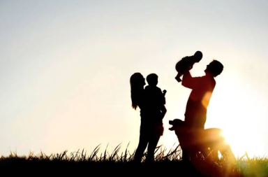 Silhuetas escuras de um casal feliz com os seus dois filhos pequenos e um cão num campo aberto de relva, celebrando a harmonia familiar sob a forte e radiosa luz dourada do pôr do sol, refletindo a atmosfera pacífica estudada na teosofia.